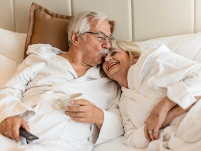 An older man and woman lie in bed together both wearing white bathrobes. She rests her head on his shoulder and laughs as he kisses the bridge of her nose.