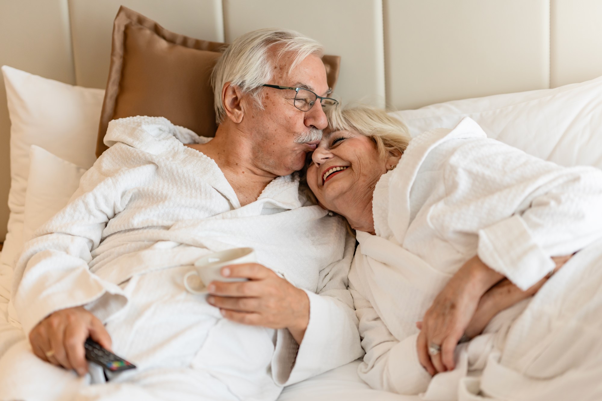 An older man and woman lie in bed together both wearing white bathrobes. She rests her head on his shoulder and laughs as he kisses the bridge of her nose.