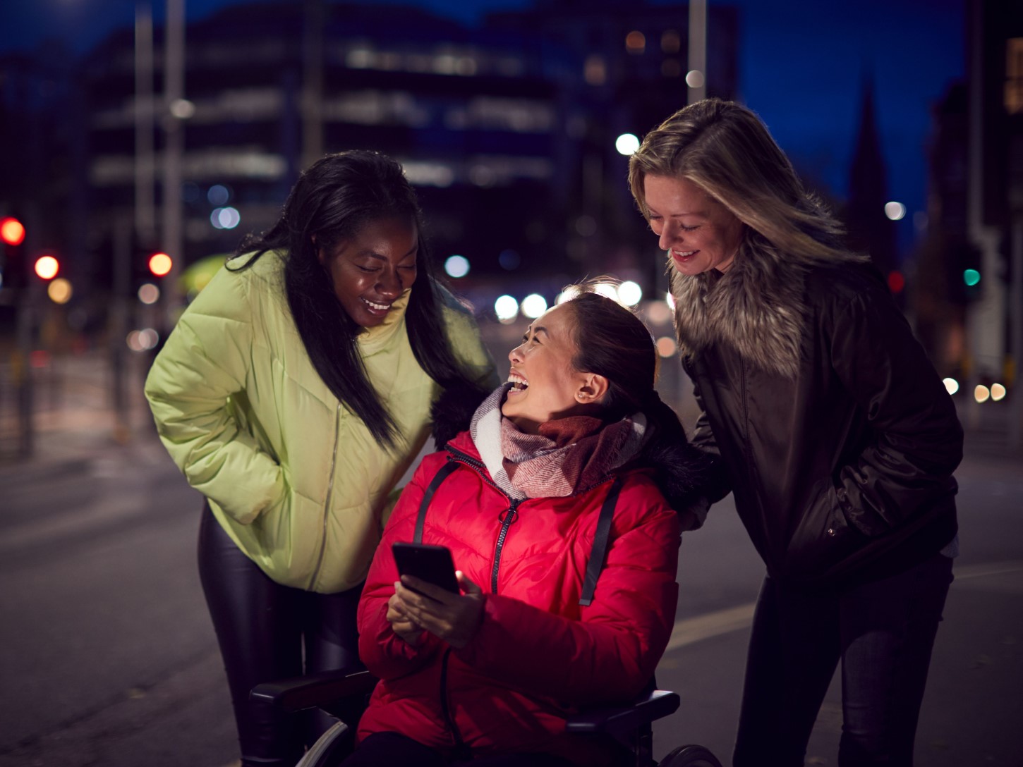 Three women out at night, one woman in a wheelchair shows them a picture on her phone that makes them all laugh.