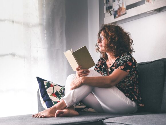 Image of a woman sitting on a bed with an open book staring out the window.