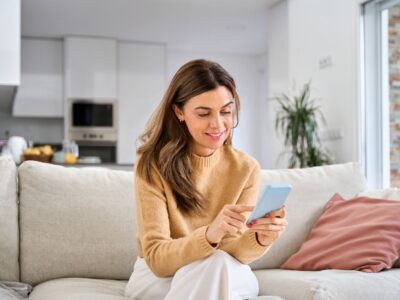 A woman sits on a sofa looking at a phone, smiling at what she reads.