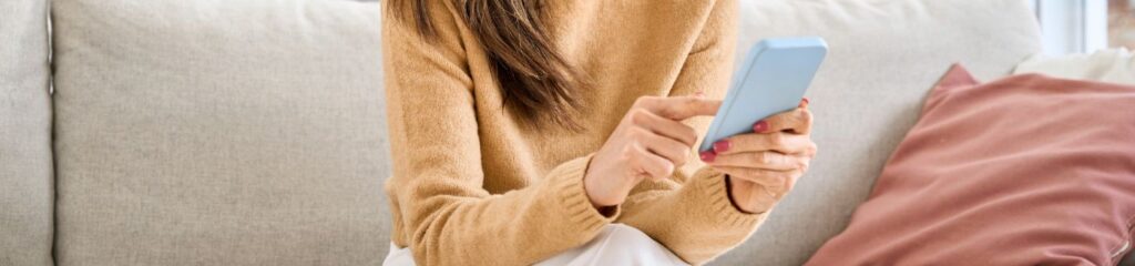 A woman sits on a sofa looking at a phone, smiling at what she reads.