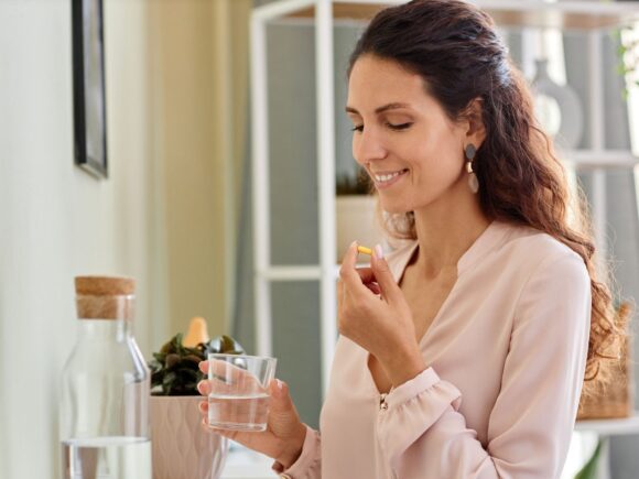 Photo of a lady looking down at her hands. She is holding a glass of water in one hand and a vitamin in the other.