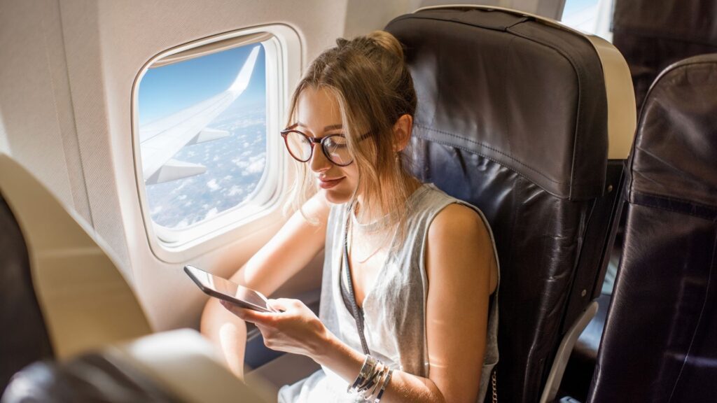 Photo of a lady sat on an aeroplane using her phone. She is sat next to the window with the window blind open showing the plane wing and clouds.