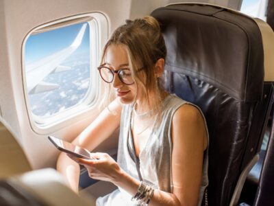 Photo of a lady sat on an aeroplane using her phone. She is sat next to the window with the window blind open showing the plane wing and clouds.