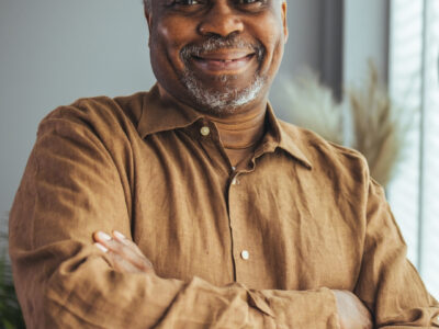 Photo of a elderly man looking towards the camera smiling with his arms crossed.