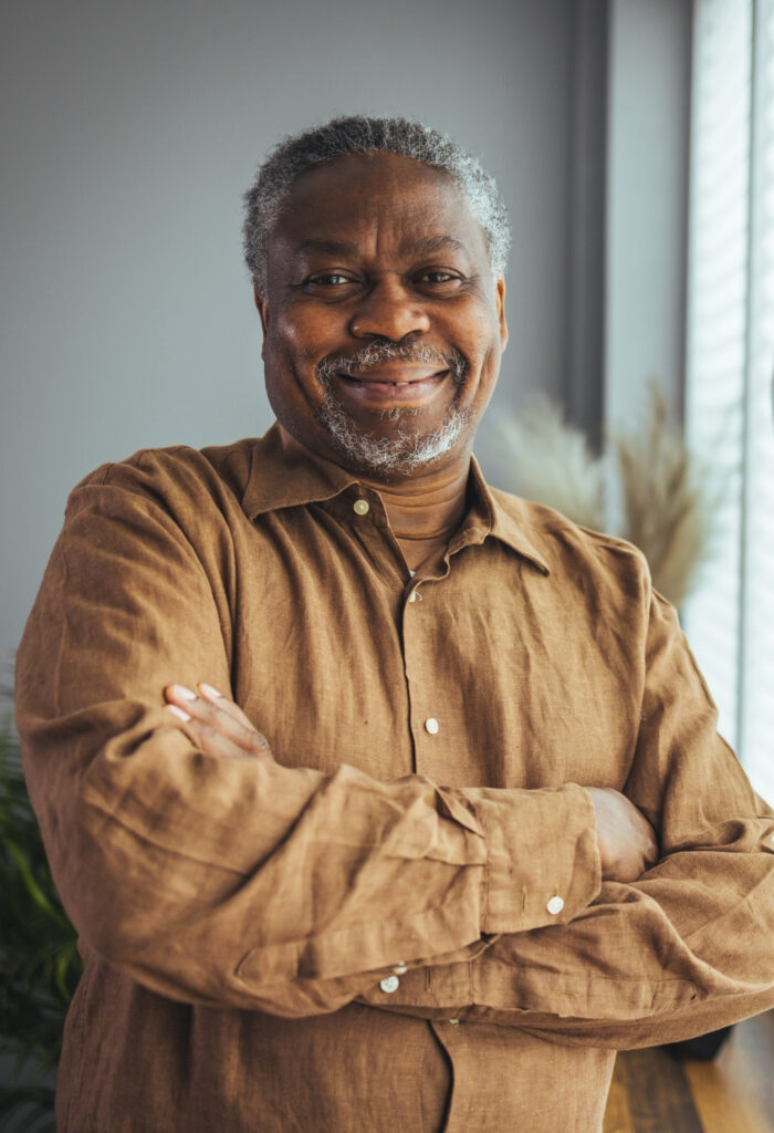 Photo of a elderly man looking towards the camera smiling with his arms crossed.