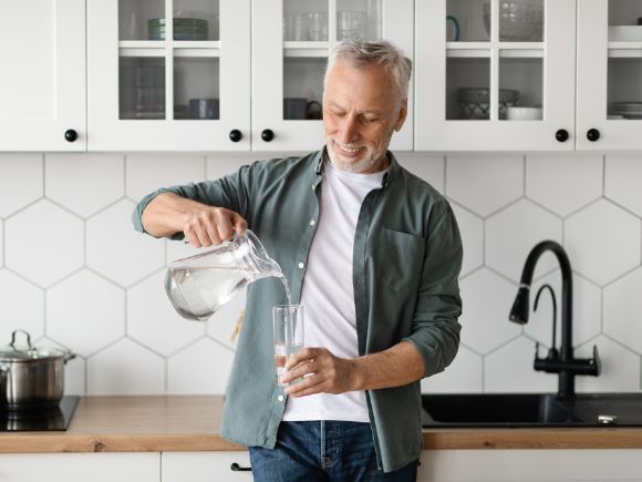 A man stands in a kitchen leaning against a worktop while pouring water from a jug into a glass.