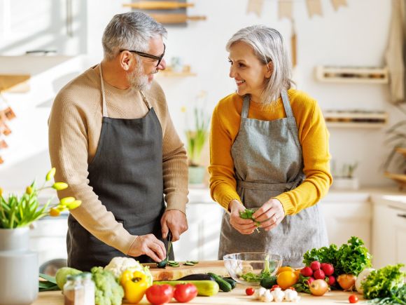 An older couple smile as they talk to each other and prepare vegetables on a kitchen island.