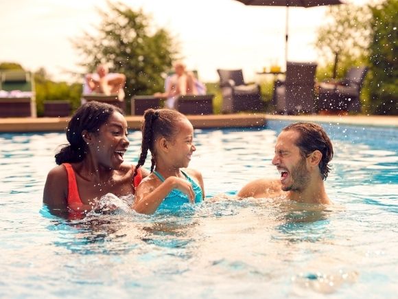 A couple play in an outdoor swimming pool with their child, the little girl splashes her father, they are all smiling.