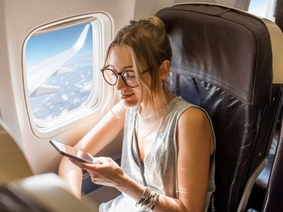 Photo of a lady sat on an aeroplane using her phone. She is sat next to the window with the window blind open showing the plane wing and clouds.