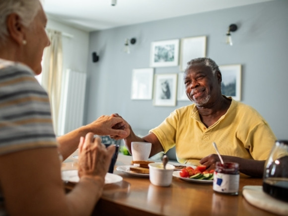 Photo of an elderly couple sitting down at a table. The wife is holding her husbands hand across the table.