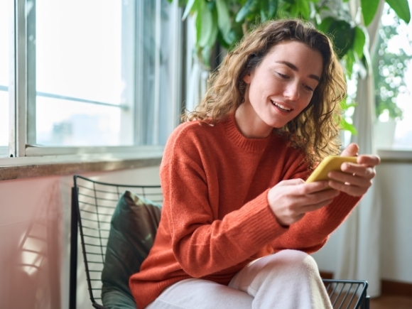 Photo of a young woman sat on a chair looking down at her mobile phone smiling