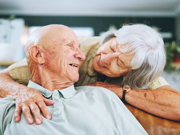 Image of an elderly man sat on a sofa, his elderly partner is holding his chest from behind. They are looking towards each other smiling.